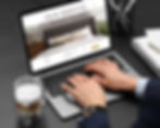 Hands typing on a laptop showing "Online Reservation" page with a bedroom image. Latte, notebook, and pen on a dark table. 