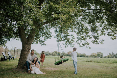 a father caresses his pregnant wife while leaning against a tree while his 3 other children play in a saucer swing under the tree