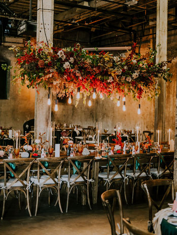 Indoor reception with a suspended floral chandelier over long wooden tables.