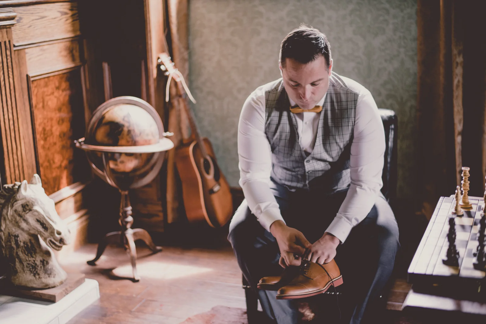 a man sits on a chair with his bowtie and vest while putting on his dress shoes