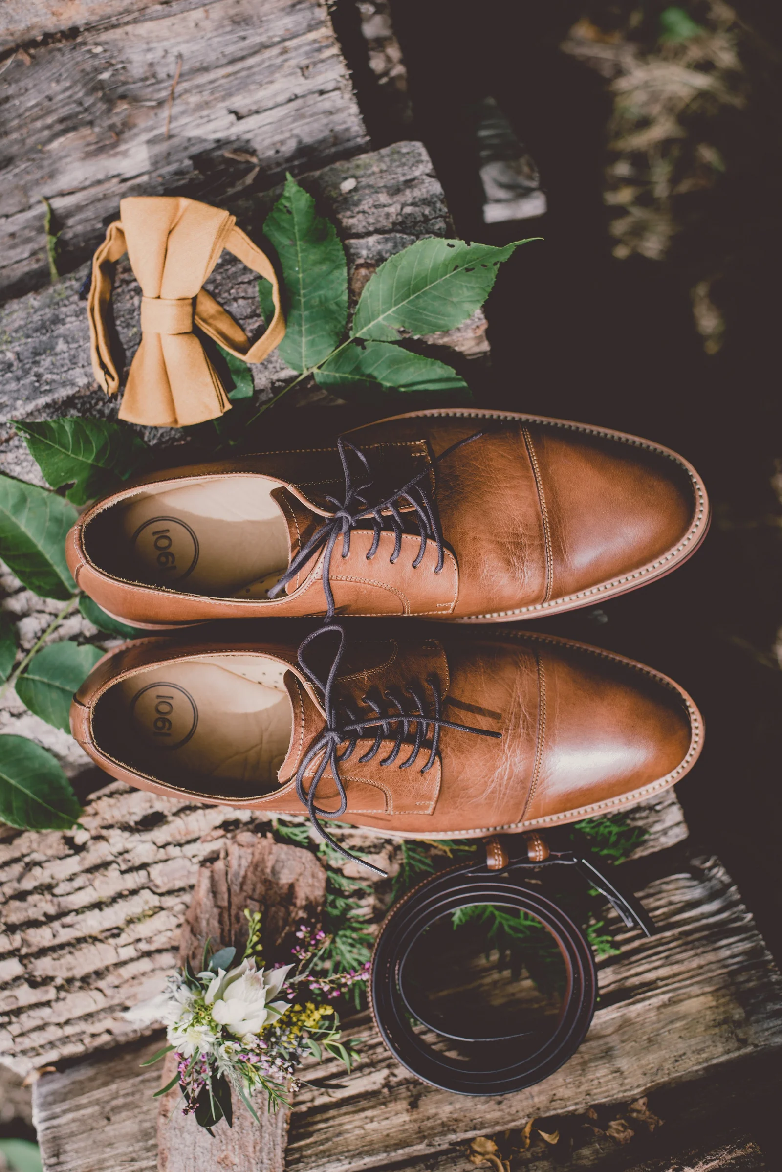an arrangement of men's dress shoes, belt and gold bow tie sit on wood