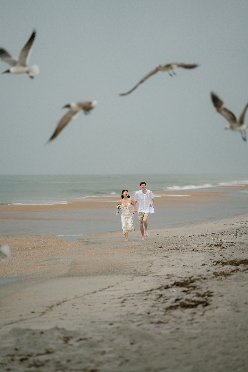 Couple in beach attire joyfully runs on a sandy beach as seagulls fly above. The ocean and cloudy sky form the serene background.