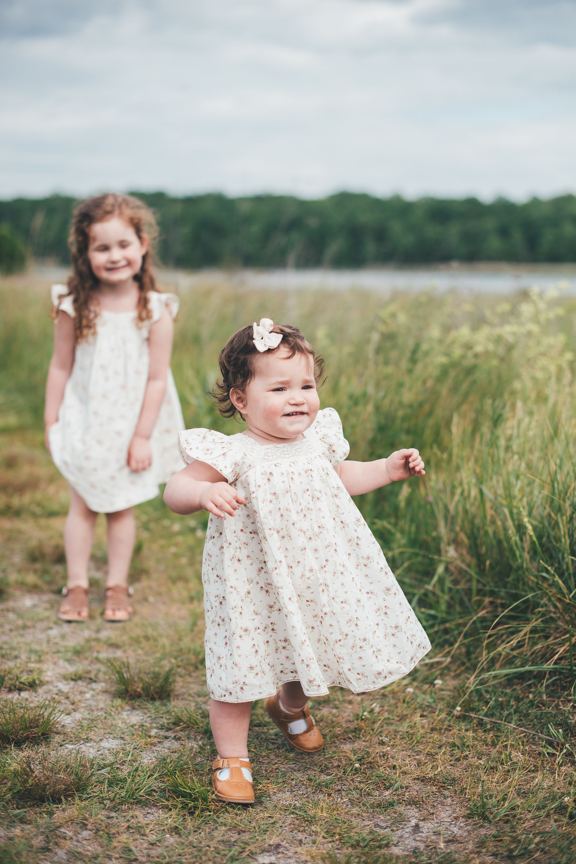 Toddler girl in white floral dress runs down a field path in Hardwick NJ while her older sister watches