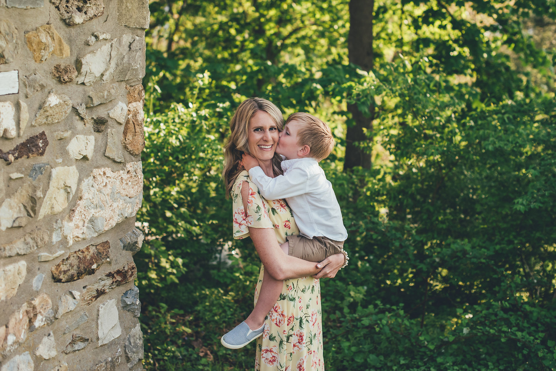 Photo session photo of mother in spring yellow pastel floral dress holding son in front of stone house in Valley Forge PA