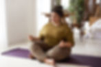 Woman meditating cross-legged on a purple mat in a cozy living room. Wearing olive top and brown pants, eyes closed, calm ambiance.