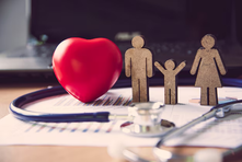 Wooden family figures, stethoscope, and red heart on documents. Background shows a blurred laptop. Mood is caring and health-focused.