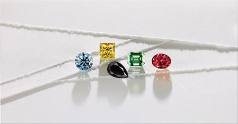 A vibrant array of fancy color diamonds from Mount Tisé, showcasing a blue round diamond, a yellow radiant diamond, a green emerald-cut diamond, a black pear-shaped diamond, and a red oval diamond, arranged elegantly on a soft white surface.