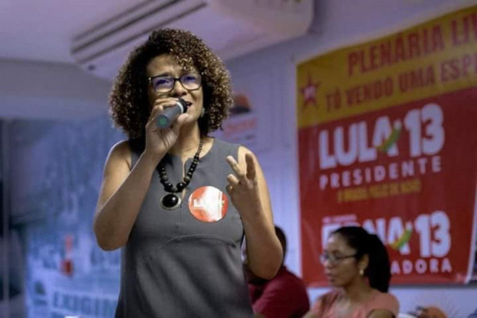 Edna Sampaio falando ao microfone usando um vestido cinza e um adesivo vermelho 'LULA 13' na roupa.