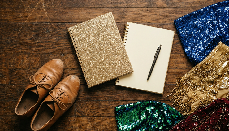 Overhead view of a glittering rehearsal notebook with dance shoes and costume fabric on a wooden floor