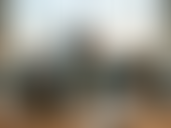 Ein realistisches Foto, das eine überforderte junge asiatische Frau an ihrem überfüllten Schreibtisch in einem modernen Großraumbüro zeigt. Im Hintergrund, durch eine große Glasfront bei Abenddämmerung, ist der Kölner Dom zu sehen. Die Frau hält in ihrer linken Hand ein Smartphone, das ein Anrufsymbol und einen Anrufernamen anzeigt, und balanciert in ihrer rechten Hand einen offenen Laptop mit einem Team-Meeting-Bildschirm. Zwei weitere Laptops, ein zweites Smartphone, zwei geöffnete Notizbücher mit handschriftlichen Notizen (sowohl Englisch als auch Deutsch), Stifte, zwei Kaffeetassen (eine mit dem Logo einer lokalen Kölner Rösterei), eine Schale Müsli und Pflanzen sind auf dem Schreibtisch verteilt. Ihr Gesichtsausdruck ist besorgt und gestresst, während andere Kollegen im Hintergrund weiterarbeiten. Die Szene ist eine visuelle Metapher für Multi-Tasking und Arbeitsstress.