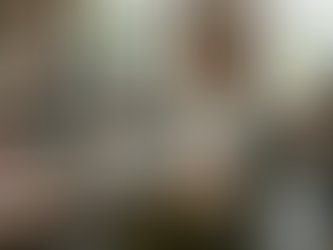 Eine junge Frau mit rötlich-braunen Locken sitzt mit geschlossenen Augen an einem großen Holztisch in einem hellen Gemeinschaftsbüro (Coworking Space). Sie trägt ein stylisches Outfit bestehend aus einem gemusterten Blazer, einem beigen Rollkragenpullover und einem grünen Rock mit Blumenmuster. Ihre Hände hält sie sanft auf ihren Bauch, der von einem warmen, goldenen Lichtschein sanft erstrahlt, was das „Hören auf das Bauchgefühl“ symbolisiert. Auf dem Tisch vor ihr liegen ein Laptop, Design-Bücher (darunter eines mit dem Titel „Bauhaus“), Stoffproben und ein offenes Skizzenbuch. Im Hintergrund arbeiten weitere Personen an ihren Schreibtischen in einem modernen Industrie-Ambiente mit großen Fenstern und Ziegelwänden. Die Atmosphäre wirkt ruhig, fokussiert und intuitiv.