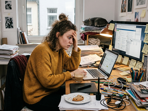 Eine junge, kreativ wirkende Frau sitzt sichtlich überfordert an einem chaotischen Schreibtisch im Homeoffice. Sie trägt einen gelben Strickpullover, hält sich mit einer Hand verzweifelt die Stirn und starrt mit geweiteten Augen auf ihren Laptop. Der Arbeitsplatz ist überladen mit Stapeln von Papier, Skizzenbüchern, unzähligen Post-its am Monitor, Stiften und einem Grafiktablett. Im Hintergrund herrscht Unordnung in den Regalen, eine Katze schläft unbeeindruckt auf einem Stapel Kleidung, und das trübe Tageslicht am Fenster unterstreicht die angespannte, stressige Atmosphäre.