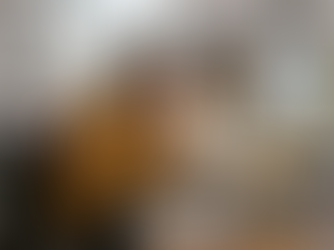 Eine junge, kreativ wirkende Frau sitzt sichtlich überfordert an einem chaotischen Schreibtisch im Homeoffice. Sie trägt einen gelben Strickpullover, hält sich mit einer Hand verzweifelt die Stirn und starrt mit geweiteten Augen auf ihren Laptop. Der Arbeitsplatz ist überladen mit Stapeln von Papier, Skizzenbüchern, unzähligen Post-its am Monitor, Stiften und einem Grafiktablett. Im Hintergrund herrscht Unordnung in den Regalen, eine Katze schläft unbeeindruckt auf einem Stapel Kleidung, und das trübe Tageslicht am Fenster unterstreicht die angespannte, stressige Atmosphäre.