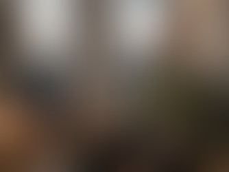 Ein moderner, stilvoll gekleideter Mann in einem olivgrünen Strickpullover sitzt aufrecht an seinem Schreibtisch im Homeoffice. Während auf seinem Laptop-Bildschirm ein Online-Meeting mit anderen Teilnehmern zu sehen ist, hat er die Augen geschlossen und macht eine bewusste Atemübung zur Entspannung. Seine Hände ruhen flach auf seinen Oberschenkeln, und sein Gesichtsausdruck ist ruhig und konzentriert. Auf dem aufgeräumten Holzschreibtisch stehen eine Sanduhr, eine Tasse Kaffee und Kopfhörer; im Hintergrund ist durch ein großes Fenster eine Stadtlandschaft bei Dämmerung zu erkennen. Die Szene vermittelt Achtsamkeit und Stressmanagement im Arbeitsalltag.