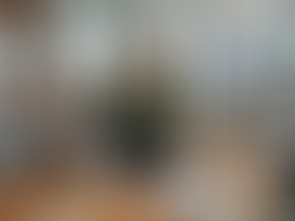 Eine surreale, humorvolle Szene in einem modernen Konferenzraum einer Kreativagentur. Ein übergewichtiger Mann in einem stylischen, moosgrünen Strickpullover, dunkler Jogginghose und weißen Sneakern sitzt im Lotussitz mitten auf dem großen Holz-Konferenztisch und meditiert mit geschlossenen Augen. Er wirkt vollkommen ruhig, während das Meeting um ihn herum ungestört weiterläuft. Fünf weitere diverse Teammitglieder sitzen am Tisch: Ein Mann links arbeitet am Laptop, eine ältere Frau neben ihm schaut auf ihren Bildschirm, und auf der rechten Seite diskutieren eine Frau und ein Mann, während sie Notizen machen. Im Hintergrund sind Brick-Wände mit Moodboards und Text wie „CLIENT PITCH“ sowie ein großer Monitor mit einer Video-Konferenz zu sehen. Der Raum ist hell und voller Arbeitsmaterialien, was den Kontrast zur meditativen Stille des Mannes auf dem Tisch betont.