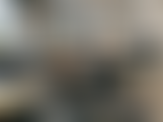 Ein surreal wirkendes Szenario in einem modernen Großraumbüro im Industrie-Stil: Ein junger Mann mit Brille und Strohhut liegt entspannt auf einer grauen Sonnenliege, die mitten im Gang platziert ist. Er trägt ein stylisches Outfit aus Sakko, grünem Strickpullover, dunkler Jeans und braunen Lederboots. Mit einem Lächeln trinkt er durch einen Strohhalm aus einer echten Kokosnuss. Während er die Ruhe genießt, herrscht um ihn herum geschäftiger Büroalltag: Kollegen sitzen an Schreibtischen vor Monitoren oder stehen in Gruppen zusammen. Durch die großen Fenster im Hintergrund ist eine gelbe Straßenbahn und die Silhouette des Kölner Doms zu sehen. Im Vordergrund liegen Design-Bücher mit Titeln wie „BAUHAUS“ und „DEEP WORK“, was den Kontrast zwischen seiner extremen Entspannung und der produktiven Umgebung unterstreicht.