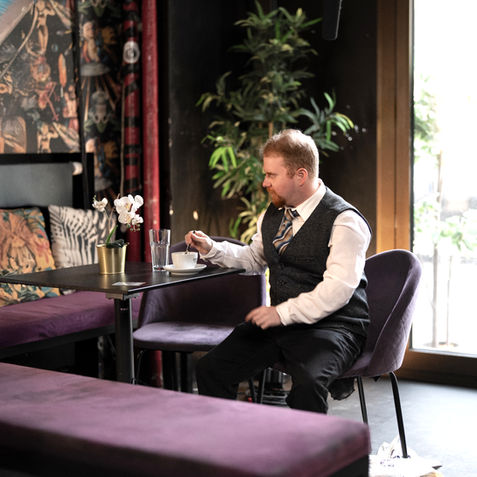 „Schauspieler Christopher Seban als Charakterdarsteller am Set von Camera Acting München: Ein Mann mit rötlichem Bart im grauen Westenesemble sitzt nachdenklich an einem Cafétisch in der Münchner Altstadt.“