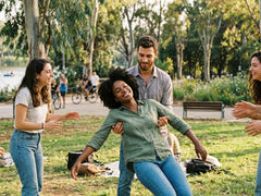 קבוצת חברים צעירים מבצעים 'תרגיל אמון' (Trust Fall) בפארק ציבורי חיצוני שטוף שמש. במרכז התמונה, אישה שחורה עם שיער אפרו, לבושה בחולצה ירוקה וג'ינס, נופלת לאחור בעיניים עצומות. גבר מזוקן תופס אותה מאחור, בעוד שלושה חברים נוספים עומדים מסביב ומחייכים. כולם מחייכים ושמחים, מוקפים בתיקים על הדשא.