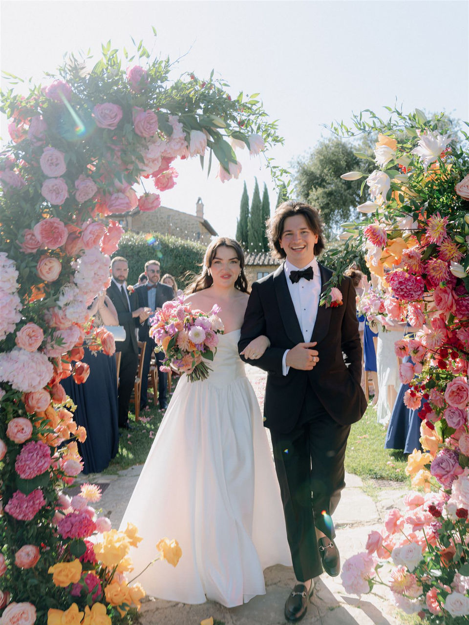 wedding couple walking through colorful floral arch during ceremony