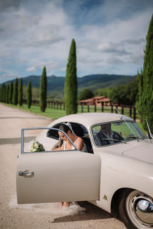 Bride stepping out of a vintage car among cypress trees in the Tuscan countryside.