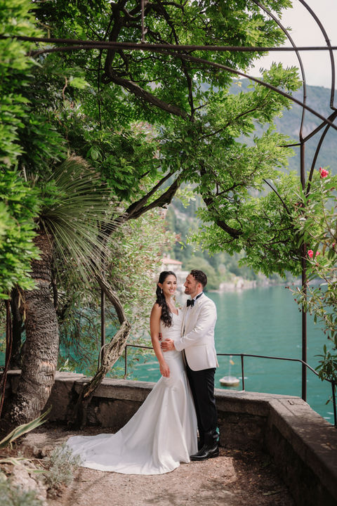 Smiling bride and groom under arch by tranquil turquoise lake.