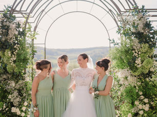 Bride with bridesmaids in sage green dresses under floral arch with Chianti hills in the background luxury wedding Tuscany