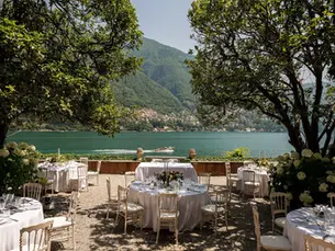 Elegant lakeside banquet tables under trees, with a boat on blue water and green mountains beyond.