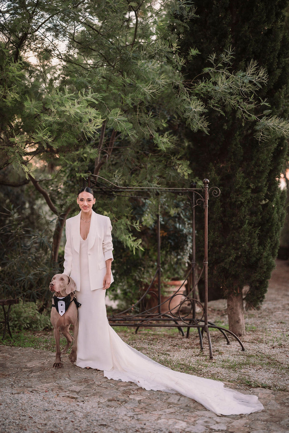 Sposa elegante in abito bianco con cane, tra alberi verdi.
