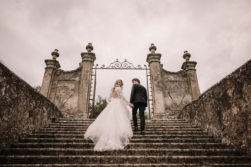 Bride and groom ascend grand stone steps towards ornate gate.