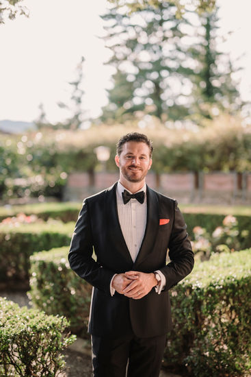 The groom in a black tuxedo smiling under the garden light of Castello di Vicchiomaggio.