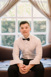Groom in formal wear sitting on couch, window backdrop, luxury wedding at Ayana Resort Bali.