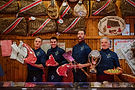Quattro macellai con tagli di carne, un trofeo e salumi appesi.