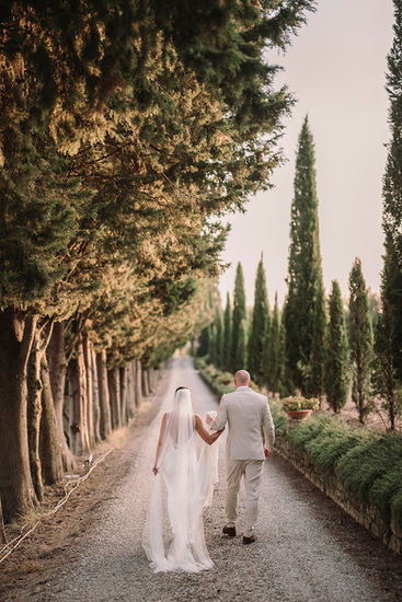 Bride and groom walking down a cypress-lined lane, veil flowing.