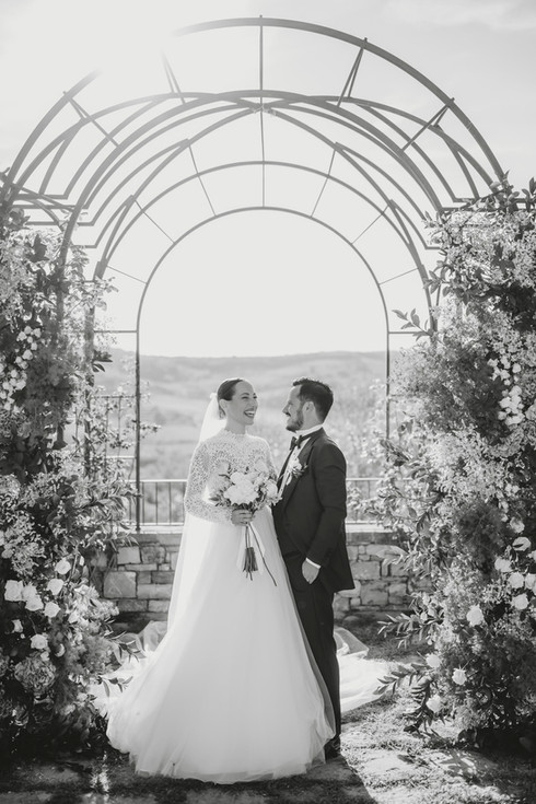Bride and groom standing beneath a floral wedding arch, sharing a natural and joyful moment
