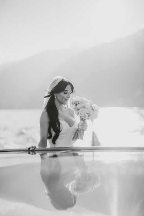 Smiling bride in wedding dress holding white bouquet, reflected in water.