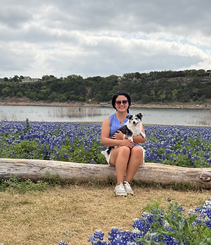 A woman holds a black and white terrier while sitting on a log with Texas blue bonnets and a lake in the background