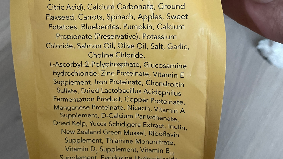 Yellow pet food bag with ingredients list including chicken, sardine, brown rice, and more. Hand holding the bag. Brand name "LOTUS" visible.