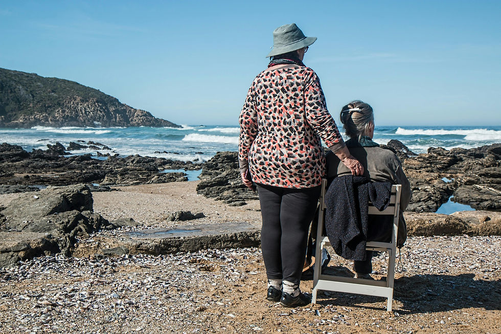 An elderly woman sitting on a chair at an empty beach, looking out at the sea. She is accompanied by another lady, standing solicitously beside her.