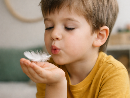 Enfant d’environ 5 à 6 ans, assis à l’intérieur, soufflant doucement sur une plume qu’il tient dans sa main. L’enfant est concentré et calme, illustrant un exercice de respiration ludique favorisant l’apaisement et la gestion des émotions.