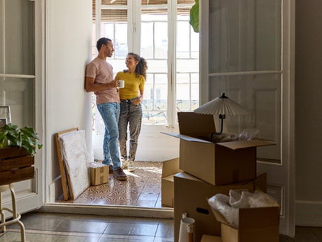 A couple stands together in a room cluttered with boxes, illuminated by light from a nearby window.