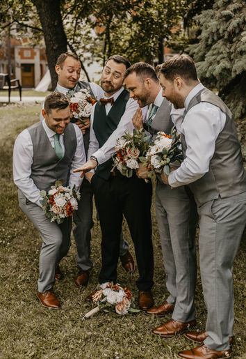 The groom proudly displays his wedding ring to his groomsmen, all of whom are gathered around him with smiles and laughter. The groom, dressed in a sharp tuxedo, holds his hand up to showcase the ring, while his groomsmen, dressed in matching suits, look on with admiration and excitement. The moment captures a joyful, lighthearted exchange between close friends before the ceremony