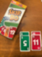 Skip-Bo card game with an open box on a wooden table. Green and red cards visible with numbers 5, 6, 11, and 12.