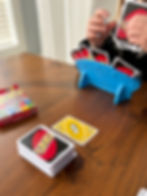 Child playing UNO at a wooden table, using a blue card holder. The table has a deck, yellow reverse card, and box. Room is softly lit.