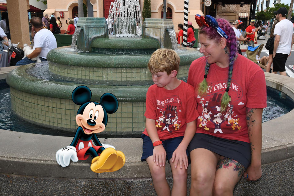 Boy and woman sit near fountain with Mickey Mouse, wearing red shirts.