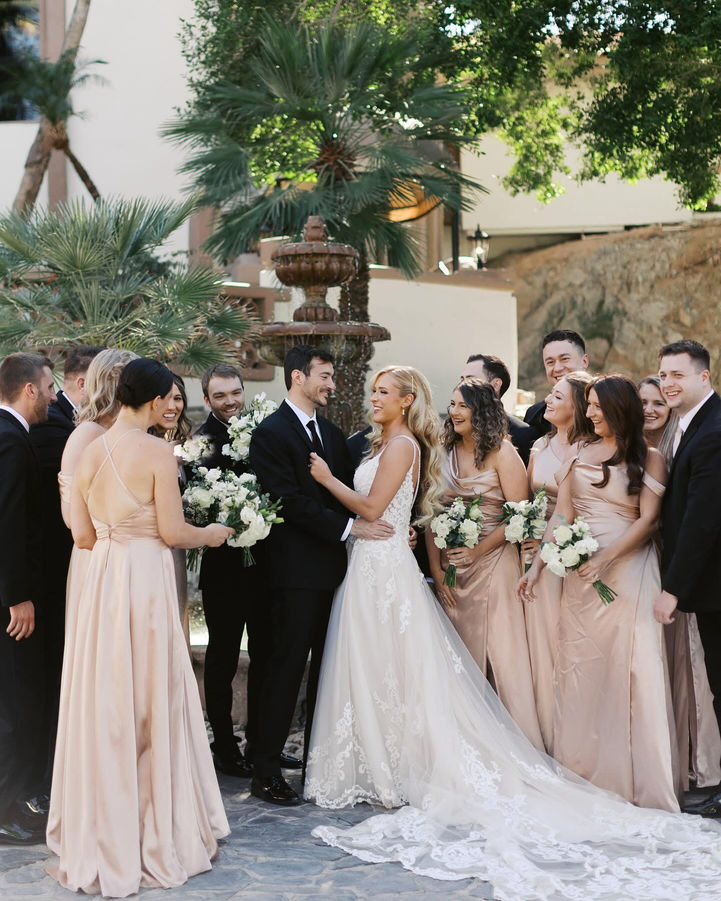 Bride and groom surrounded by full wedding party in black tuxedos and champagne dresses near a courtyard fountain