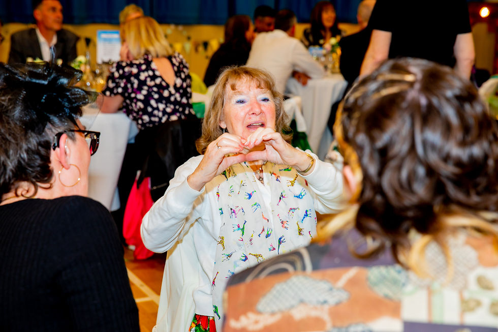 an older lady sitting at a wedding reception, she is holding her hands up  to make a heart shape at the guests infront of her