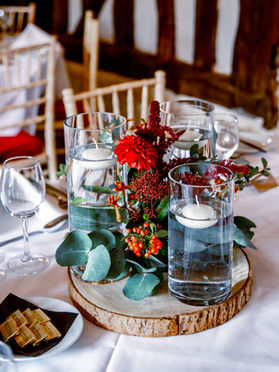 A wedding table topper that has a wooden log as the base, with red flowers and greenery and tall cylinder vases filled with water with candles floating on the top