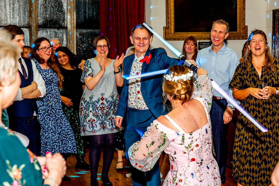 A joyful indoor gathering with people watching two adults playfully duel with toy lightsabers. Brightly colored outfits and laughter abound.
