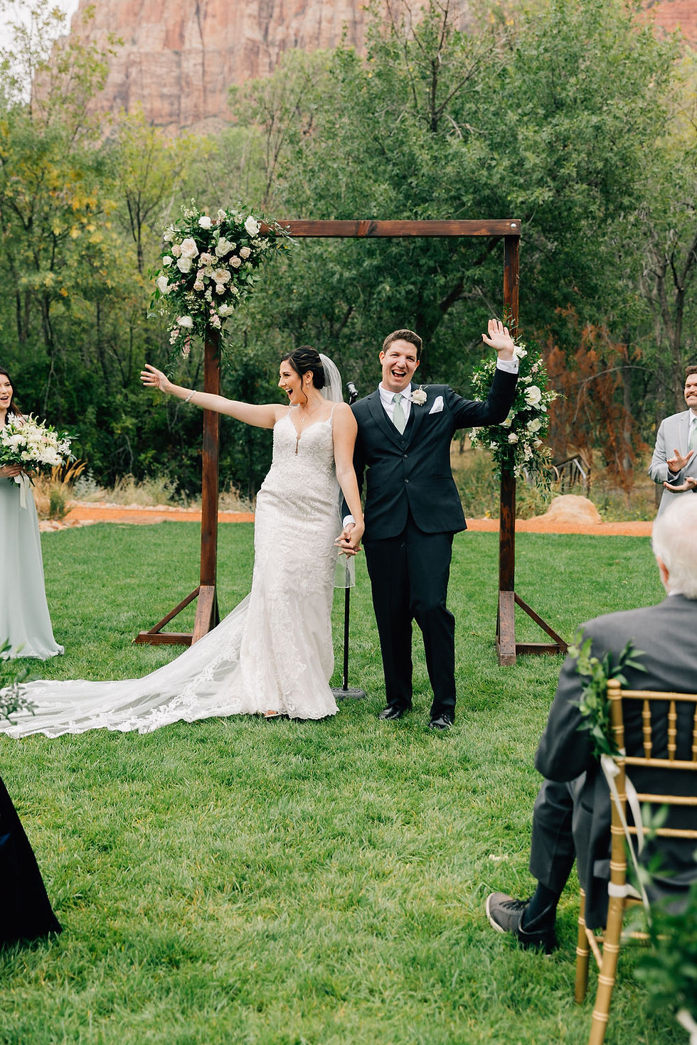 Bride in embellished wedding gown holding hands with groom in black suit with sage green tie in front of rustic boho wedding arch waving at guests as they walk down the aisle. #wedding #utahwedding #zionwedding #bohowedding #destinationwedding #southernutahwedding #eventcoordintor #ido #intimatewedding #elopement #zionweddingplanner #utahbride #outdoorwedding #weddinginspo #weddingdayready #destinationelopement #weddingtips #weddingdetails #zionbride #bohobride #brideandgroom #destinationweddingcoordinator #sohitched #weddingday #yourbigday #walkingdowntheaisle #brdieandgroomwaving #whiteroses #sagegreenwedding #bestweddingplannerinsouthernutah #happiness
