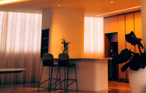 Modern kitchen with a warm glow, featuring a central island, two high chairs, and plants. Tall cabinets and sheer curtains in the background.